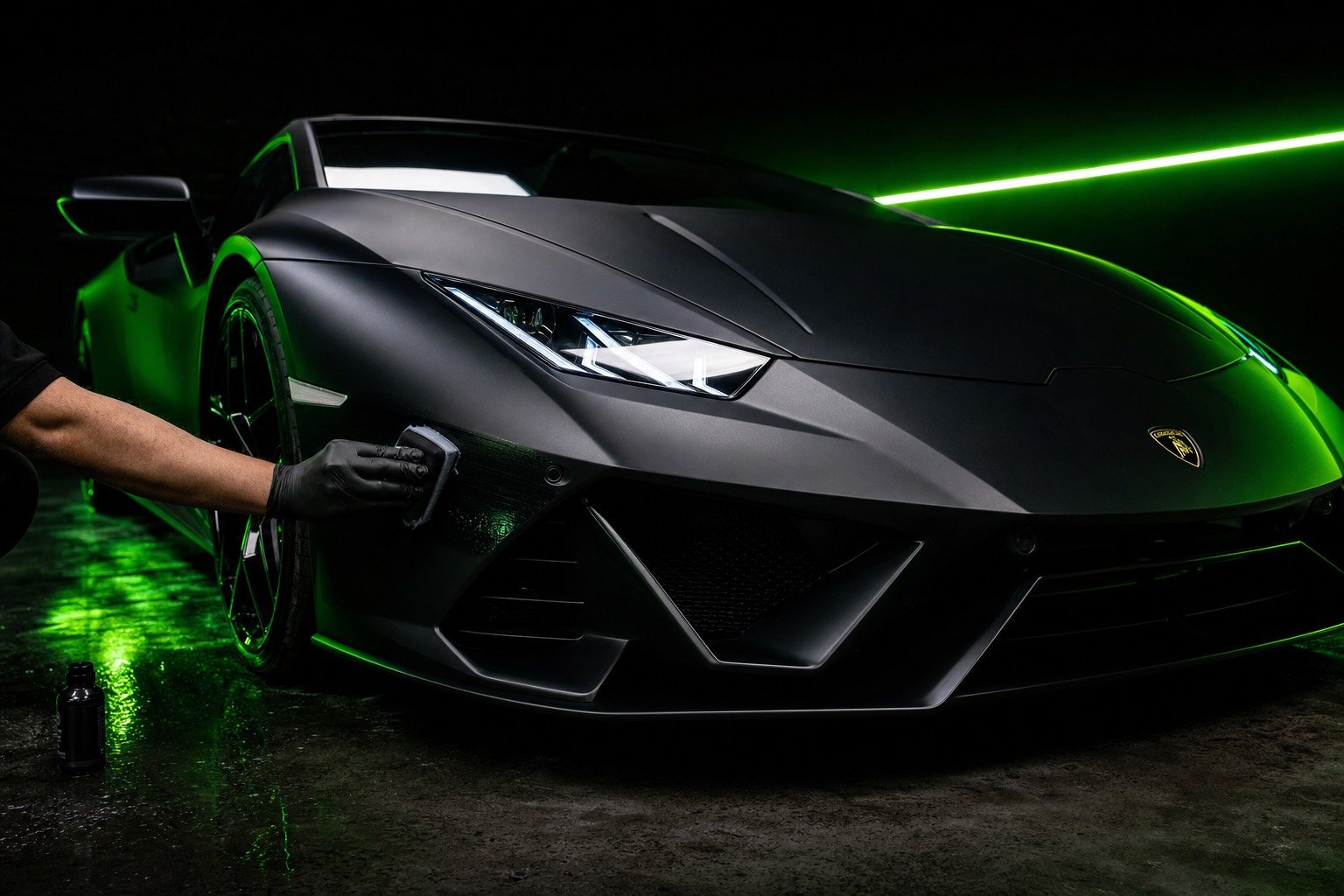A technician in black nitrile gloves applying graphene ceramic coating to the front quarter of a matte black Lamborghini Huracan under a green neon strip